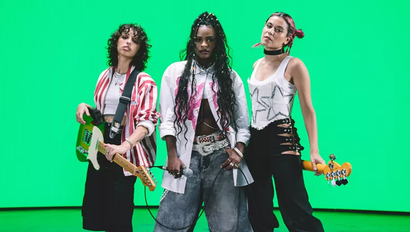 Image: Three musicians pose in front of a bright green background. The woman on the left holds a guitar and the woman on the right holds a bass behind her back.