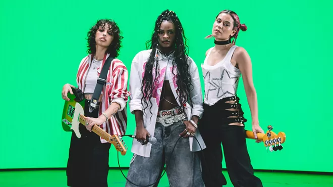 Three musicians pose in front of a bright green background. The woman on the left holds a guitar and the woman on the right holds a bass behind her back.
