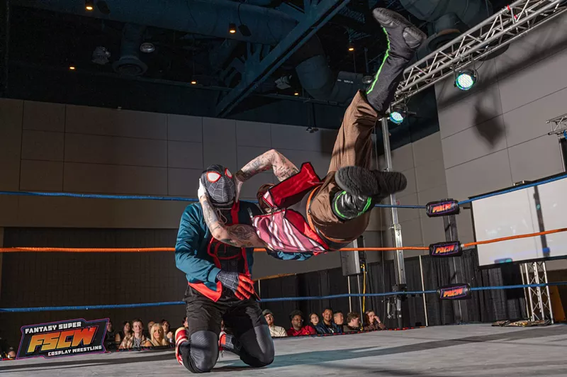 Image: two people dressed in costumes wrestle in a ring. one is suspended in the air parallel to the ground
