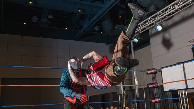 two people dressed in costumes wrestle in a ring. one is suspended in the air parallel to the ground