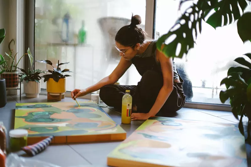 Image: color photo of Miami-based painter Orianna Montenegro at work, seated on the floor of a sunlit room with plants and a large window in the background