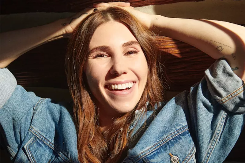 Image: close-up portrait of actress Zosia Mamet wearing a denim jacket and resting her hands on her head