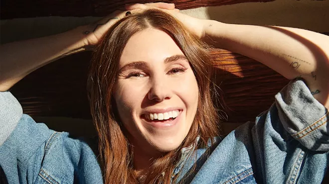 close-up portrait of actress Zosia Mamet wearing a denim jacket and resting her hands on her head