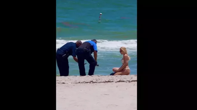 A woman in a bikini and two police officers crouch over a brick of white powder on the beach