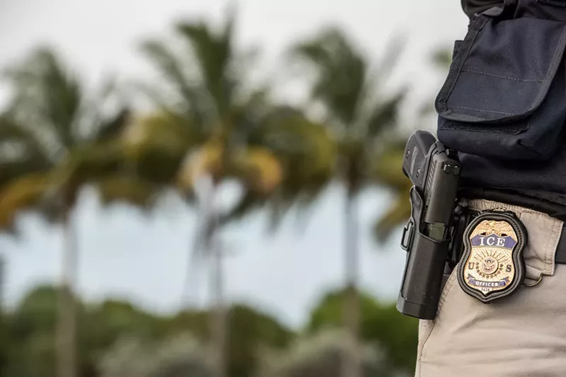 Image: An immigration officer with an ICE badge next to palm trees.