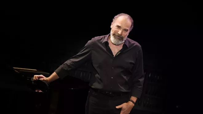 portrait of Mandy Patinkin in a black button-down shirt and pants, leaning on a black piano inside a black box theater