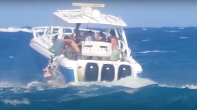 A boat full of teenagers navigates rough waters as one boy dumps a trash can full of garbage into the ocean
