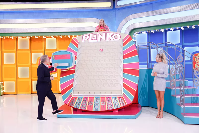 Image: A game show host and model stand on either side of a gameboard reading "Plinko." A woman stands above the gameboard