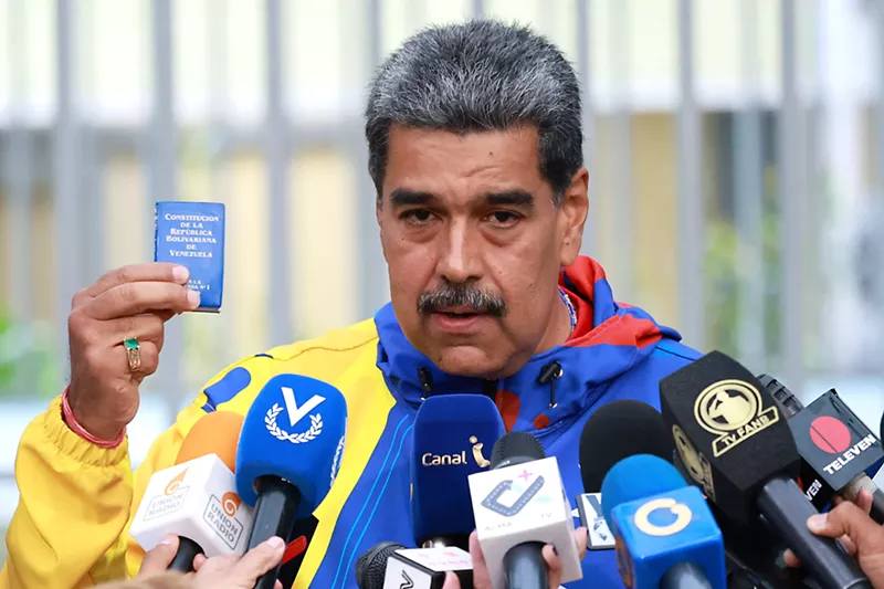 Image: Mustachioed man in front of microphones, holding a teensy copy of the Venezuelan constitution.