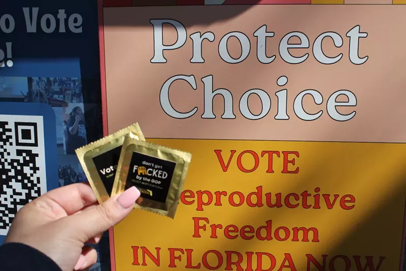 Image: A hand holds two black and gold condoms with the words “Don’t Get Fucked by the GOP. Protect Abortion in Florida" printed on the front, in front of a yellow and orange sign that reads "Protect Choice, Vote Reproductive Freedom in Florida Now."