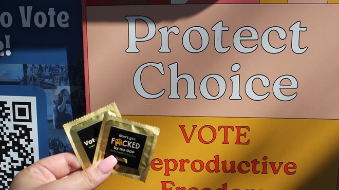 A hand holds two black and gold condoms with the words “Don’t Get Fucked by the GOP. Protect Abortion in Florida" printed on the front, in front of a yellow and orange sign that reads "Protect Choice, Vote Reproductive Freedom in Florida Now."