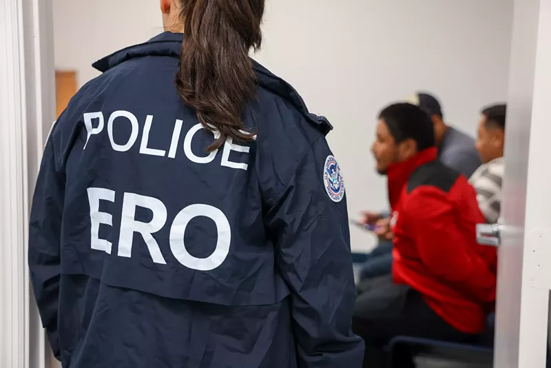 Image: A woman wearing a POLICE "ERO" jacket overlooking three men sitting in chairs blurred in the background.