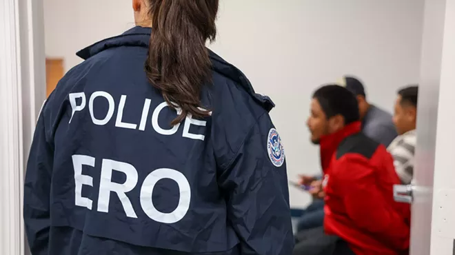A woman wearing a POLICE "ERO" jacket overlooking three men sitting in chairs blurred in the background.