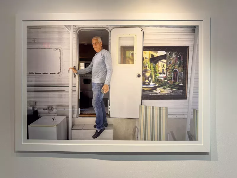 Image: A framed photo is on display in a gallery. In the image, a man stands at the entrance of his mobile home. A painting with a fountain and greenery hangs next to the door