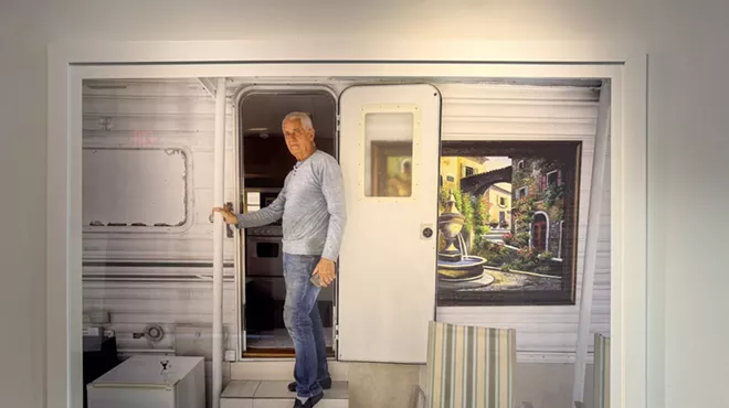 A framed photo is on display in a gallery. In the image, a man stands at the entrance of his mobile home. A painting with a fountain and greenery hangs next to the door
