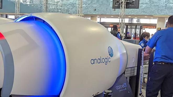 TSA agents surround a security scanning machine. A logo on the machine reads, "Analogic."