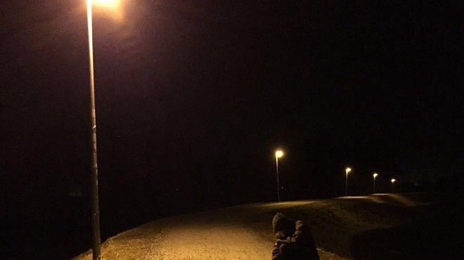 Skee Mask, face obscured, crouching down a road with a street lamp shining overhead