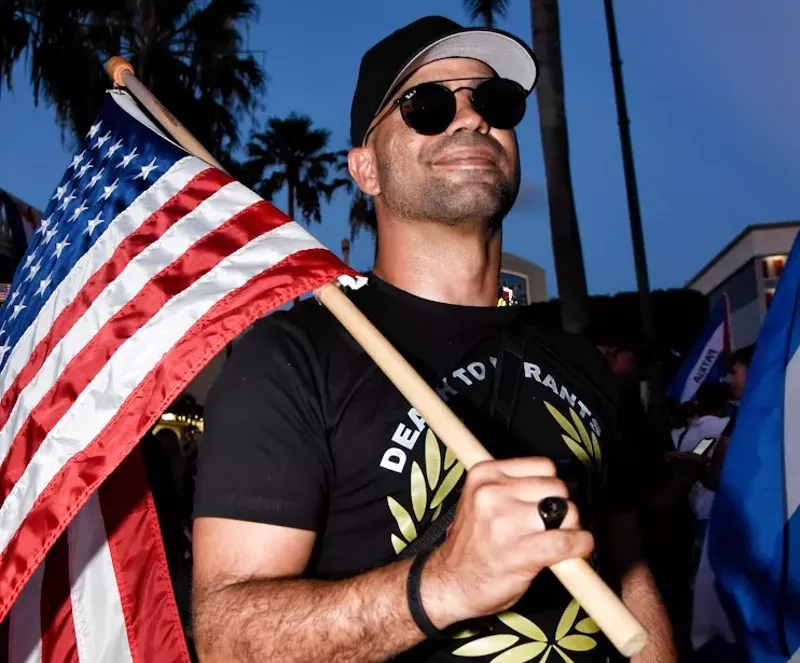 Image: photo of former Proud Boys chairman Enrique "Henry" Tarrio wearing aviator sunglasses and holding an American flag