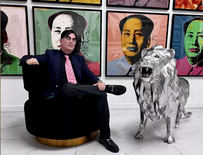 Image: photo of a seated man with a conspicuous toupee beside a large lion figurine with ostensible Andy Warhol works in the background