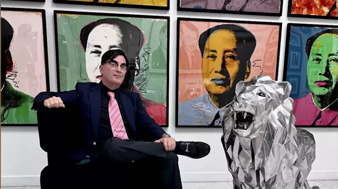 photo of a seated man with a conspicuous toupee beside a large lion figurine with ostensible Andy Warhol works in the background