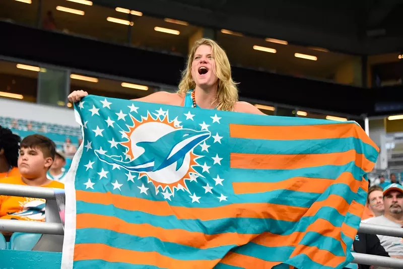 Image: A blonde woman holds a Miami Dolphins flag with teal and orange stripes on it