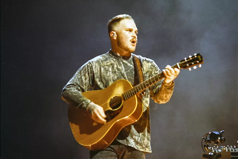 Image: Zach Bryan with his guitar in hand performing on stage