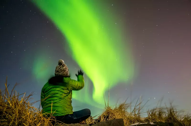 Image: A person in a beanie and thick green jacket looks up at green Northern Lights.
