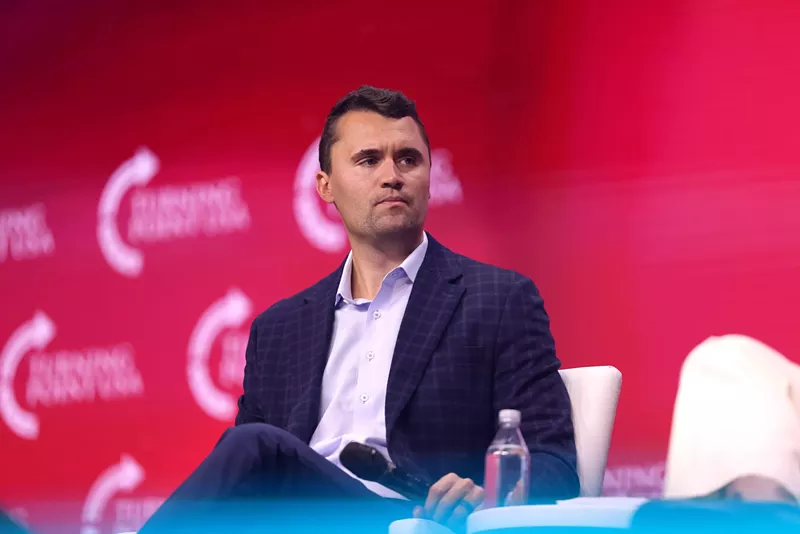 Image: color photo of a man wearing a white open collared shirt, navy blue sport coat, and navy blue pants seated at a public event against a magenta background