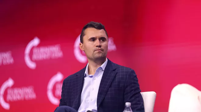color photo of a man wearing a white open collared shirt, navy blue sport coat, and navy blue pants seated at a public event against a magenta background