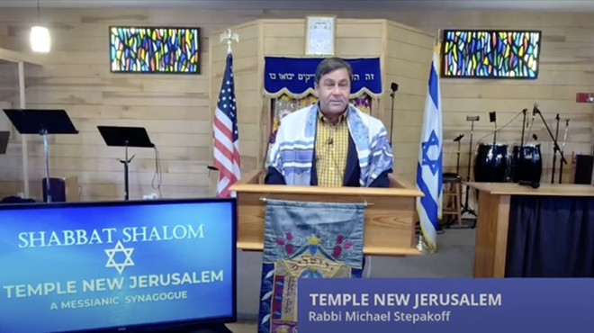 Michael Gary Stepakoff leads a shabbat service at his synagogue, Temple New Jerusalem. The 56-year-old is standing at a lectern before an Israeli and American flag and wearing a prayer shawl.