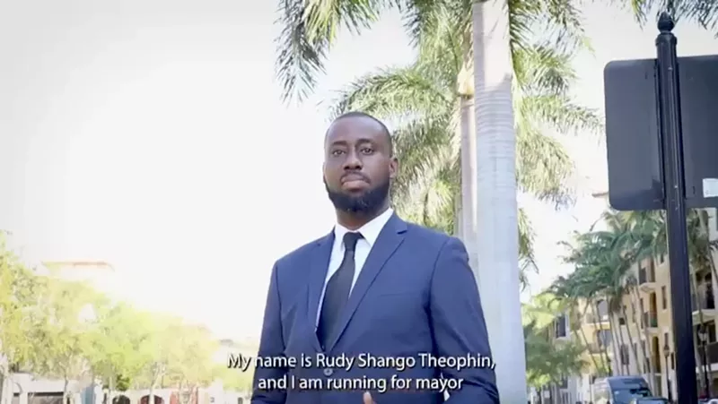 Image: video screenshot of a black man wearing a suit and tie with a caption that reads, "My name is Rudy Shango Theophin, and I am running for mayor."