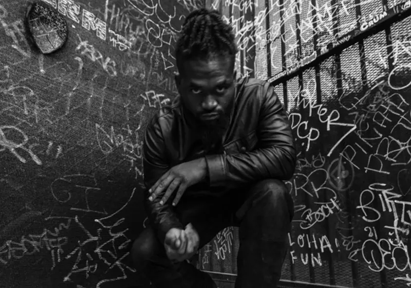 Image: black and white picture of a young man by a graffiti-covered wall.