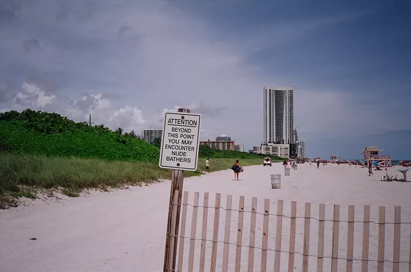 Image: a sign on the sands on the beach reads "Attention: Beyond this point you may encounter nude bathers"