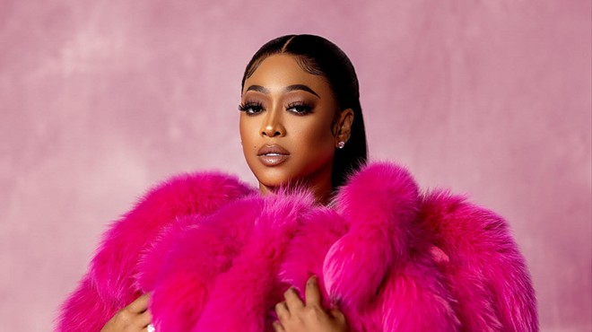 Rapper Trina wearing a pink fur coat while standing against a soft-pink background