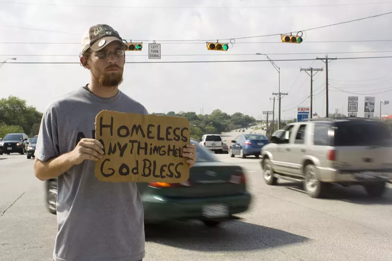 Image: A man who appears to be homeless stands on the side of a busy road holding a cardboard sign enscribed with the words: "HOMELESS, ANYTHING HELPS, GOD BLESS."