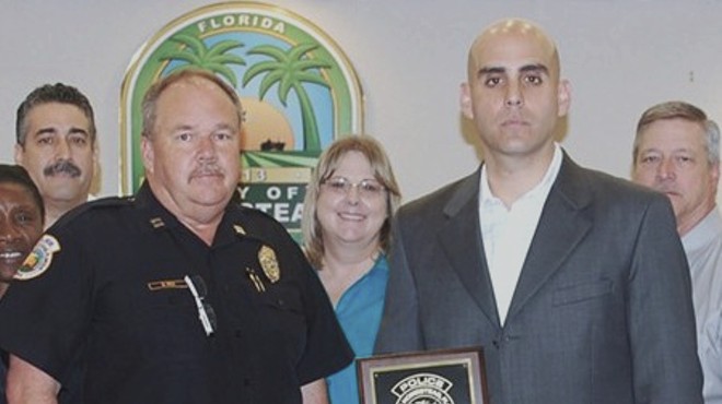 A man with a shaved head poses in plainclothes with fellow police officers, holding an "Officer of the Month" award
