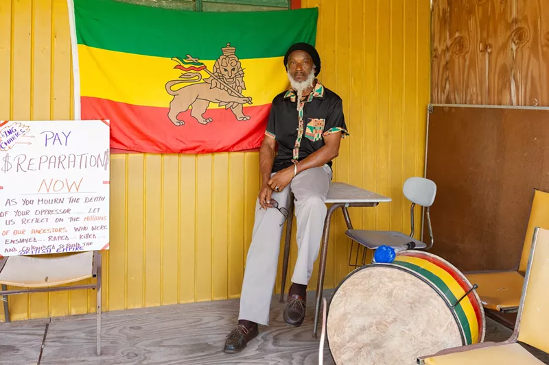 Image: Ras Iroy standing in front of the Rastafarian flag and a protest sign
