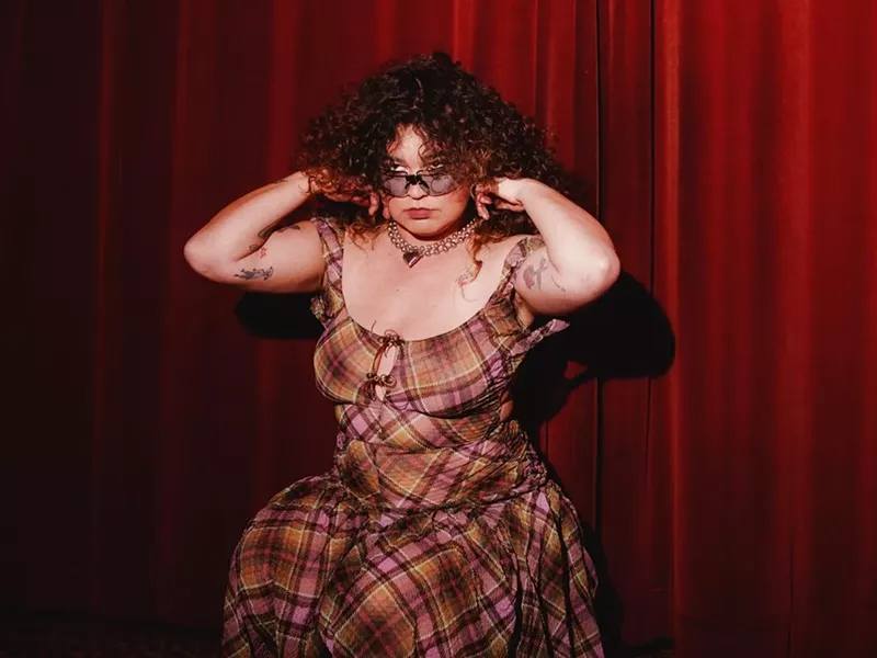 Image: color photo of singer songwriter Remi Wolf, bespectacled, clad in a low-cut plaid dress, and showily primping her hair