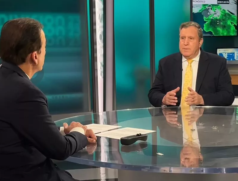 Image: photo of a well-dressed man facing the camera, sitting at a glass table across from a television anchor who's partially visible in 3/4 profile