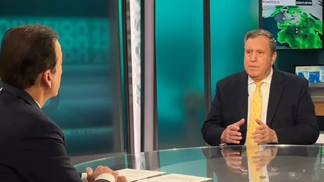 photo of a well-dressed man facing the camera, sitting at a glass table across from a television anchor who's partially visible in 3/4 profile