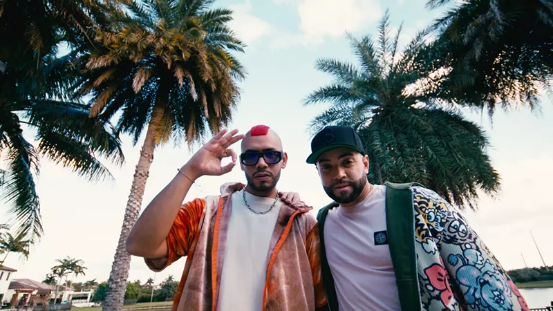 Image: Two men posing with palm trees in the background.