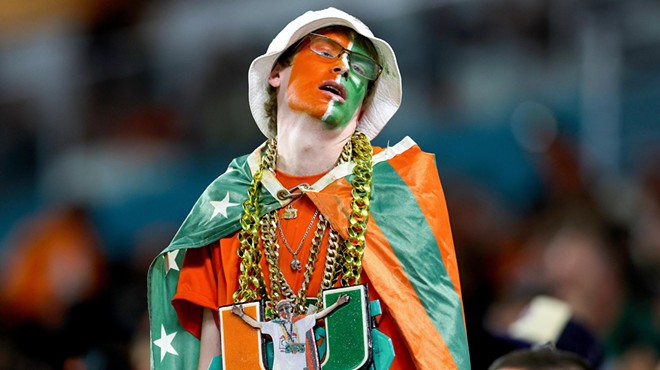 A Miami Hurricanes fanatic with his face painted is wearing gold chains and some kind of cape at a college football game