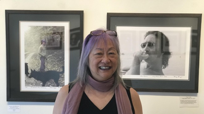 color photo of a smiling gray-haired woman in an art gallery with framed photos of John Lennon behind her