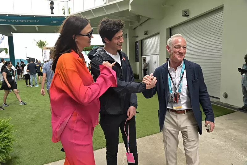 Image: A woman in a pink suit and a man with brown hair, both smiling, stand with an elderly man wearing a lanyard