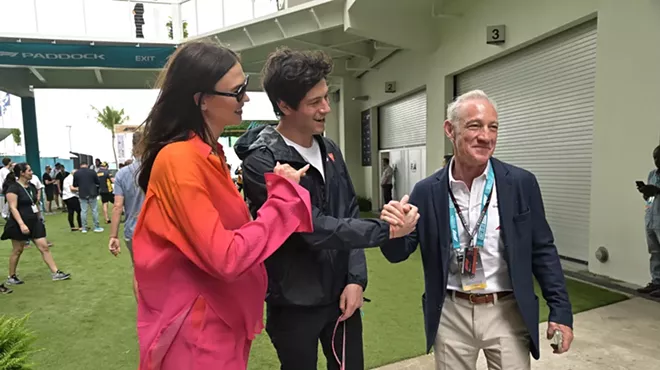 A woman in a pink suit and a man with brown hair, both smiling, stand with an elderly man wearing a lanyard