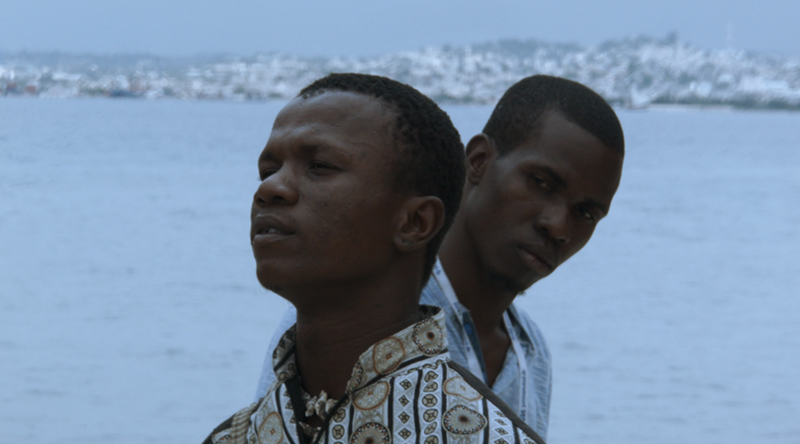 Image: a still from a film showing a close-up of a Black man standing behind another