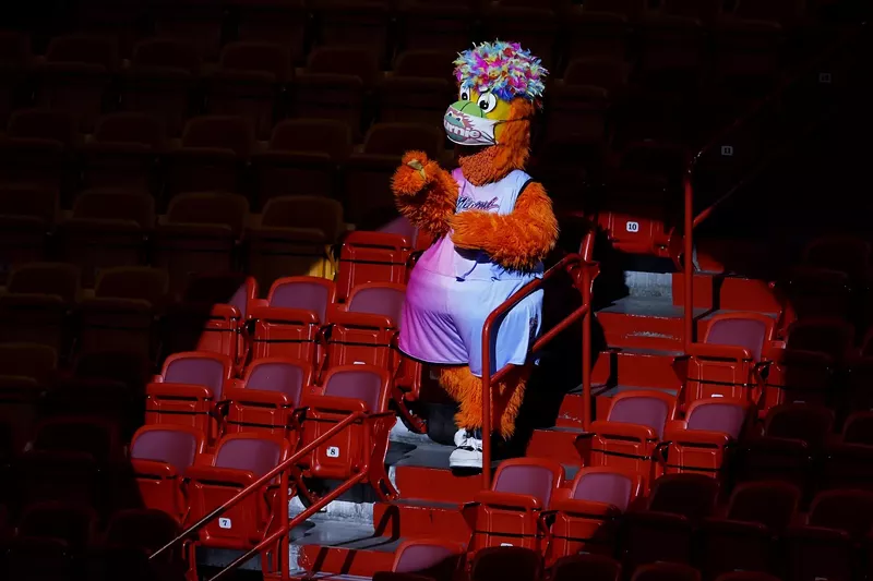 Image: photo of Miami Heat mascot Burnie against a sea of empty arena seats, wearing a Pride-ish wig