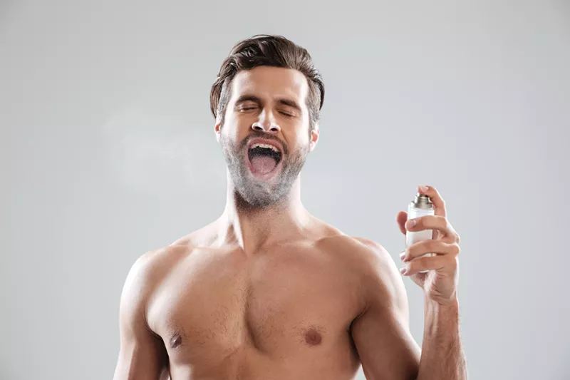 Image: A shirtless man enthusiastically spraying himself with cologne, mouth open wide, highlighting excessive fragrance habits discussed in Miami