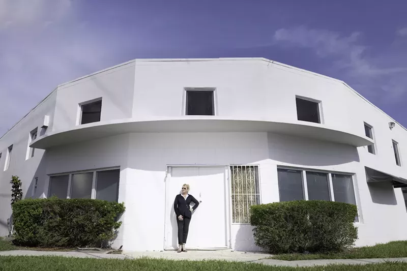 Image: photo of woman standing in the doorway of a two-story white building on a corner lot