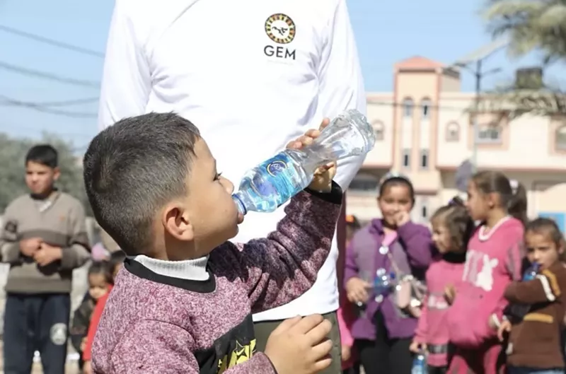 Image: A child in Gaza drinks from a water bottle in front of a person wearing a shirt with a Global Empowerment Mission logo.